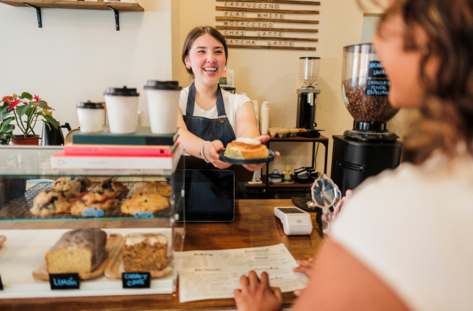 Young woman barista smiling enthusiastically, handing a freshly baked cinnamon roll on a plate to a customer at the counter of a cozy, modern cafe, providing excellent customer service