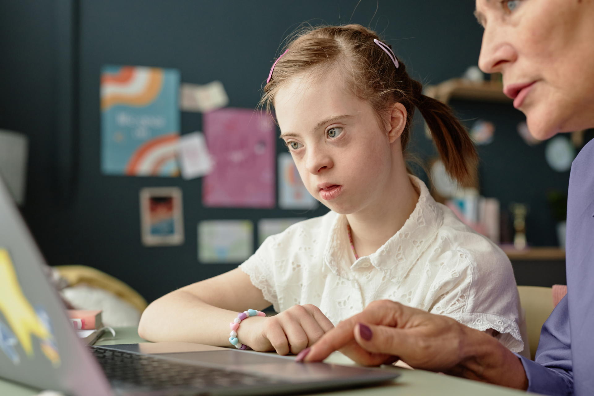Girl with Down Syndrome engaging in home learning on laptop with adult supervision in background, focusing on screen and participating in educational activity